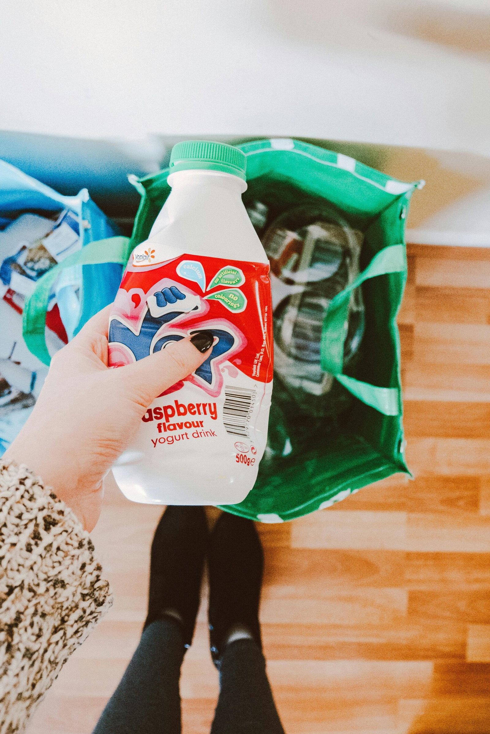 A person recycling a raspberry yogurt drink bottle into a green recycling bag.
